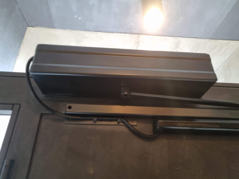 Detail instalace dveřního pohonu nastříkaného do antracitové barvy na hliníkové vstupní dveře.