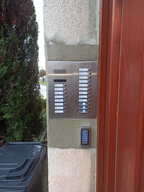 Detailní pohled na moderní digitální tablo domovního telefonu s podsvícenými jmenovkami a čtečkou s klávesnicí pro obyvatele. 