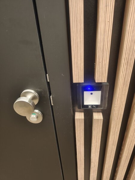 Detail instalace čtečky QR kódů pro automatické otevírání dveří na toalety (WC). Přístupový systém s elegantním zapuštěním do dřevěného obkladu umožňuje kontrolovaný vstup po naskenování platného QR kódu z účtenky nebo mobilního telefonu. Skvělé řešení pr