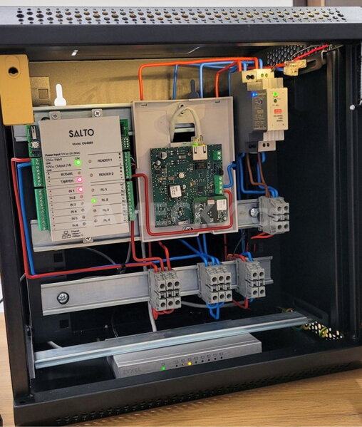 Otevřený instalační rozvaděč s profesionálně zapojenou řídící jednotkou přístupového systému Salto a síťovými prvky pro chytré otevírání dveří.