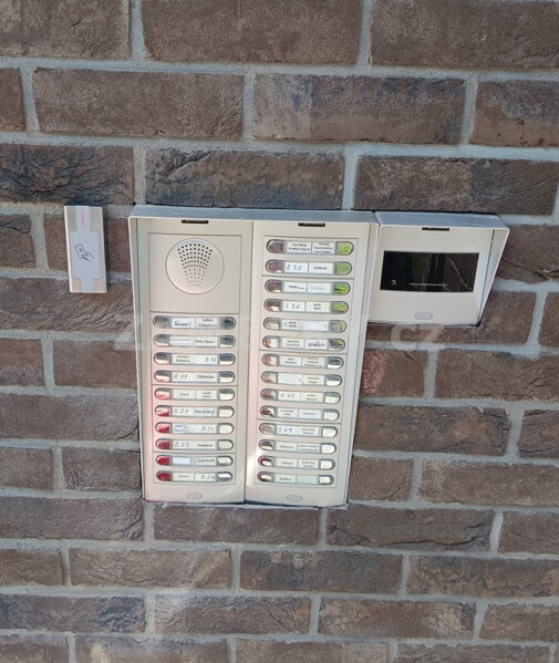 Venkovní RFID čtečka čipů a karet nainstalovaná vedle zvonkového tabla Fermax na cihlové fasádě. Spolehlivý přístupový systém pro bezpečné otevírání hlavních vchodových dveří do bytových domů (SVJ) a firemních budov.